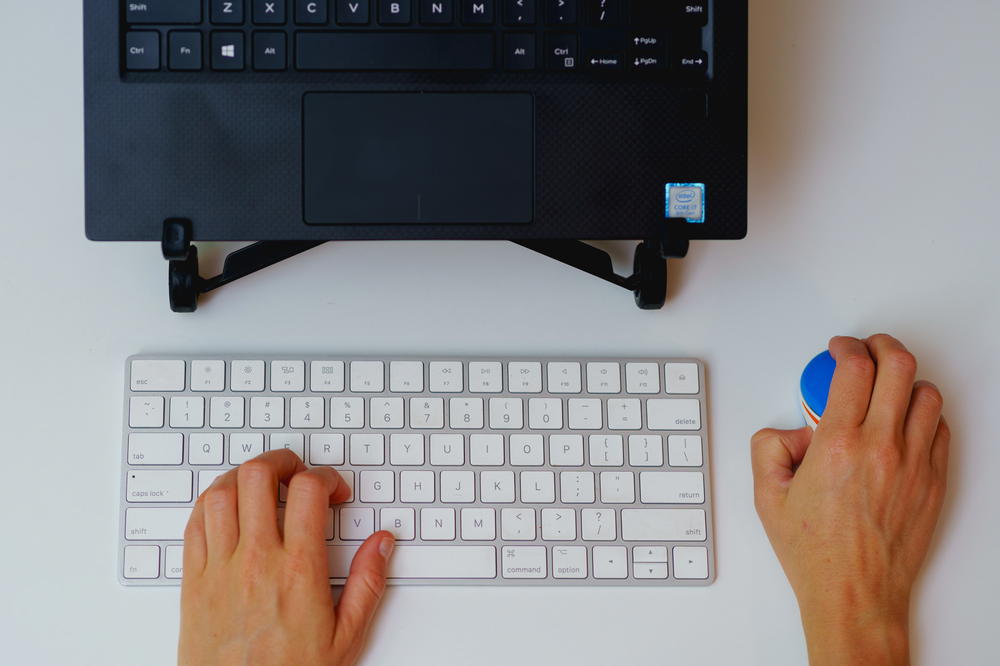 Woman using a small wireless mouse with an apple magic keyboard and laptop on a laptop stand in a home office setup