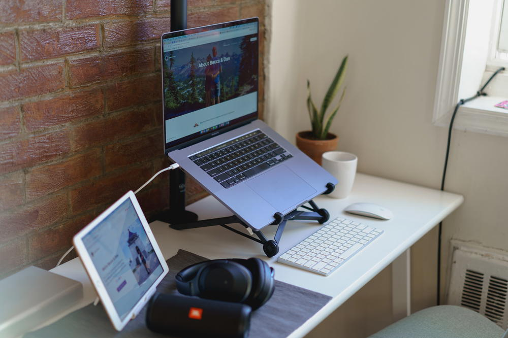 Exposed brick wall in a living room with a home office desk, silver laptop on a black laptop stand, white wireless keyboard, wireless mouse and black bluetooth head phones