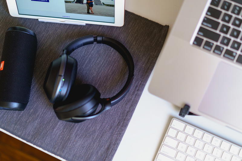 Wireless bluetooth headphones, black compact bluetooth speaker and laptop with wireless keyboard on a gray place mat on a white home office desk