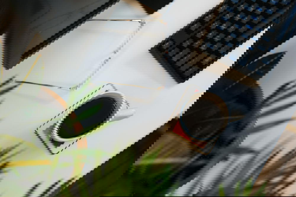 Flat lay of a home office setup with clear glasses, a white cup full of coffee, leafy green plants and a black keyboard