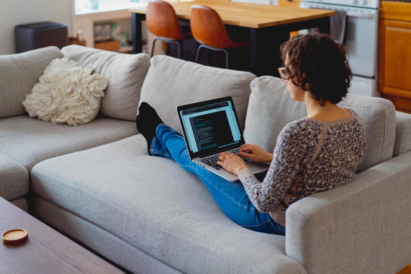 Woman sitting on a couch with a laptop working from home with a silver MacBook Pro near the kitchen
