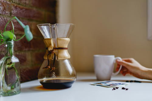 Coffee in a Chemex on a white table next to a brick wall and a hand holding a white coffee cup